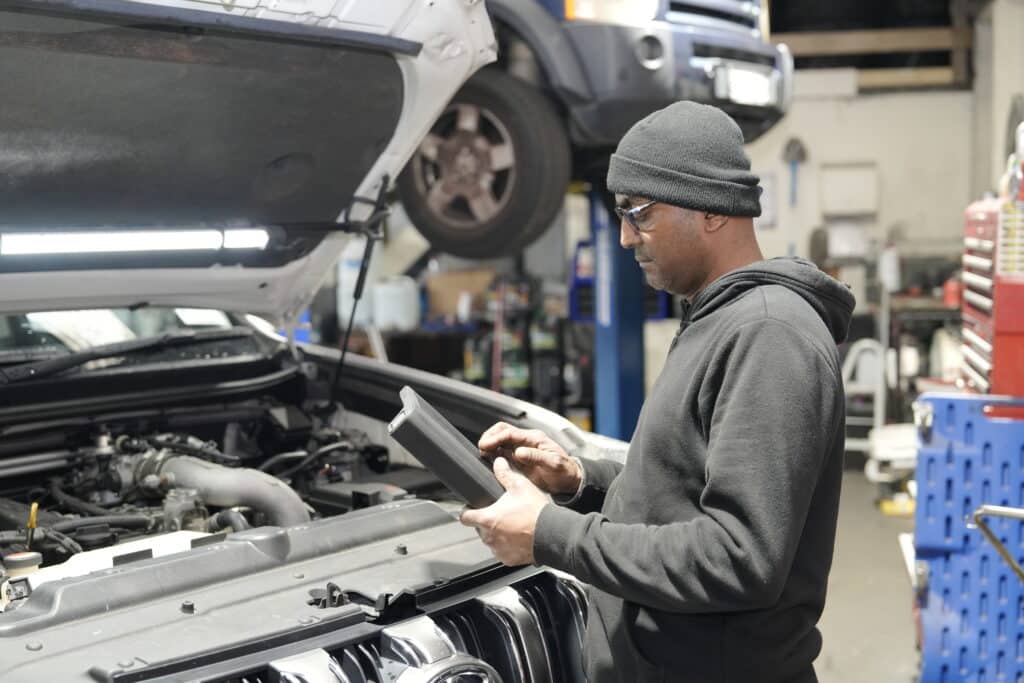 A person wearing a beanie, glasses, and a dark hoodie is working in an auto repair shop, inspecting a tablet near a white car with its hood open. Another vehicle is lifted in the background. The workshop has various tools and equipment visible.