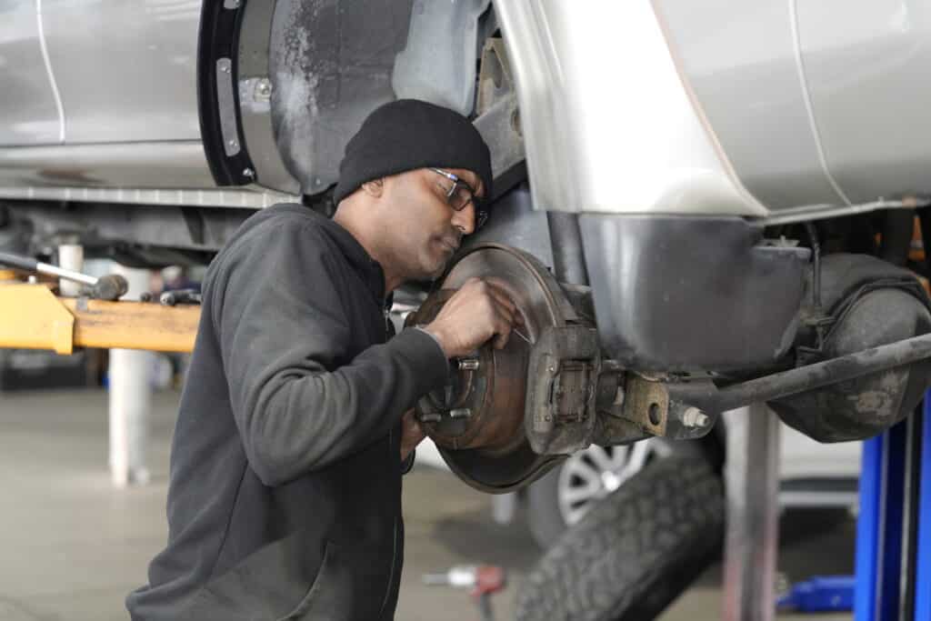 A mechanic wearing a black beanie and glasses is working on the brake system of a car elevated on a lift in an auto repair shop. He is using a tool to adjust or inspect the brake disc assembly. The background shows another vehicle and various equipment.