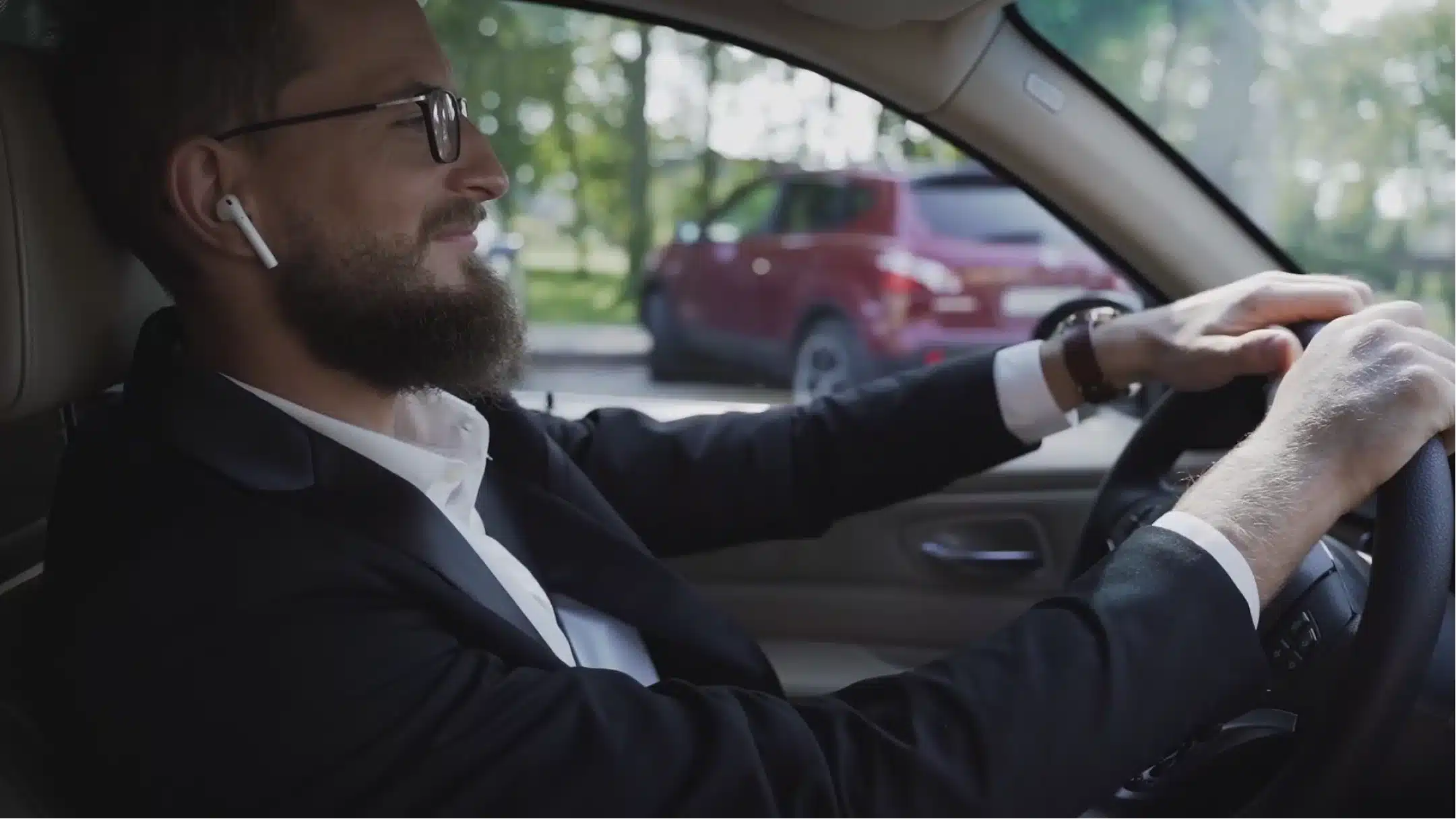 A man with a beard and glasses, wearing a black suit and white shirt, is driving a car. He is smiling slightly and looking forward. He is also wearing wireless earbuds. There is a red car and greenery visible outside the window.