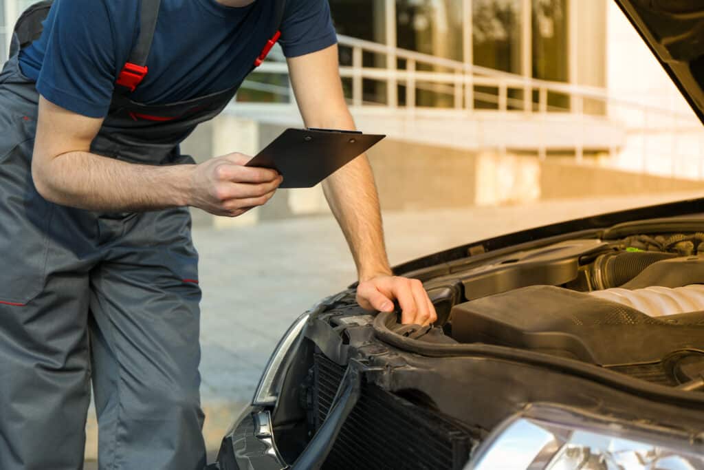 A mechanic wearing grey overalls and a navy blue t-shirt examines a car engine with the hood open. He holds a black clipboard and leans on the car, likely inspecting or diagnosing an issue in the bright daylight outside a building.