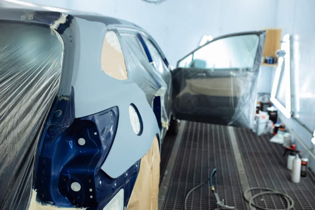 An unfinished blue car is undergoing a paint job in an auto body shop. The rear and door panels are masked with tape and plastic sheeting, and the car is inside a well-lit spray booth. The car door is open, and spray painting equipment is visible on the floor.