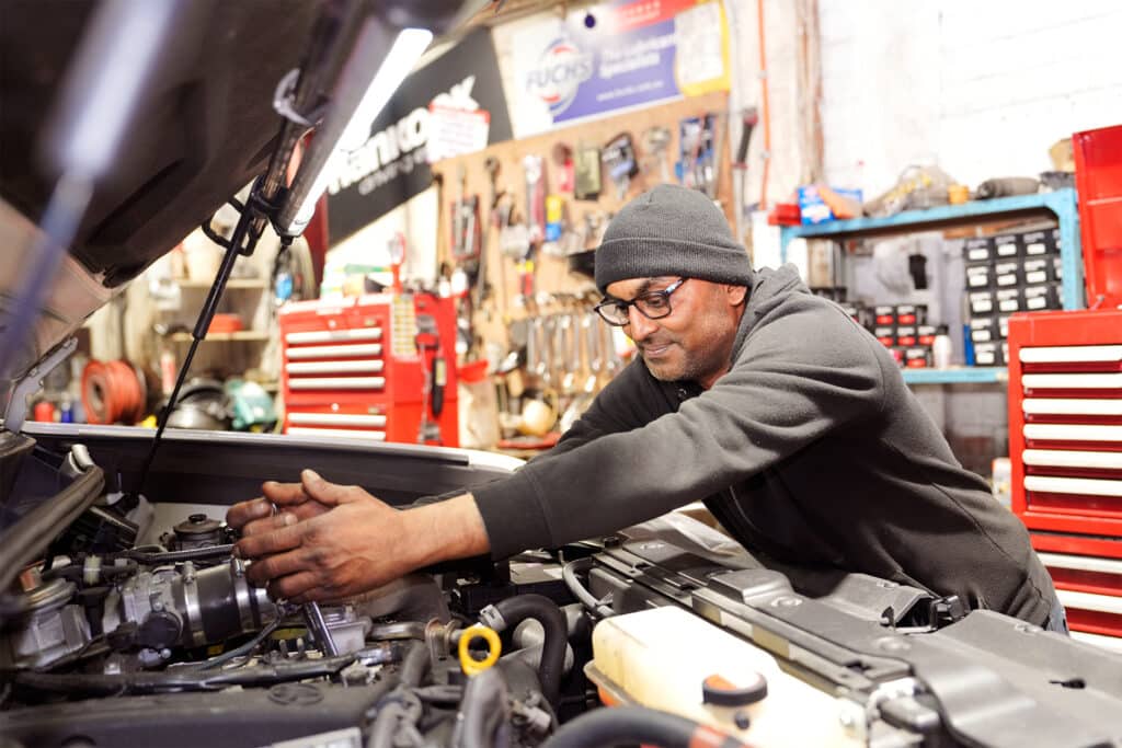 A mechanic wearing a beanie and glasses is working under the hood of a car in a garage. The workspace is filled with various tools, red tool cabinets, and automotive equipment in the background. The mechanic is focused on repairing the engine.