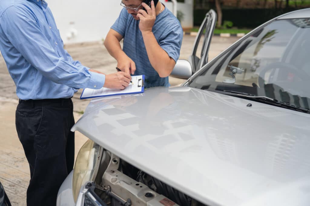 Two men stand beside a silver car with its hood open. One man in a blue shirt is making notes on a clipboard, while the other man is talking on a phone. The setting appears to be outdoors in a parking lot or similar area.