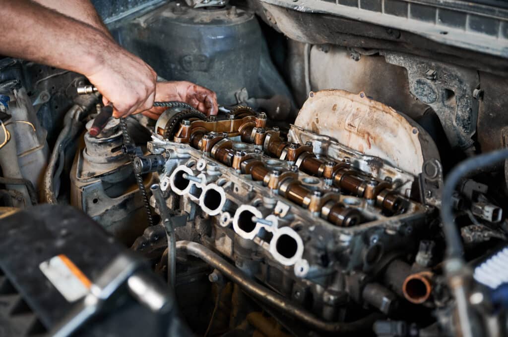 A mechanic's hands work on a disassembled car engine, focusing on the camshaft and cylinder head area. Various engine components and tools are scattered around the engine bay, indicating an ongoing repair or maintenance task.