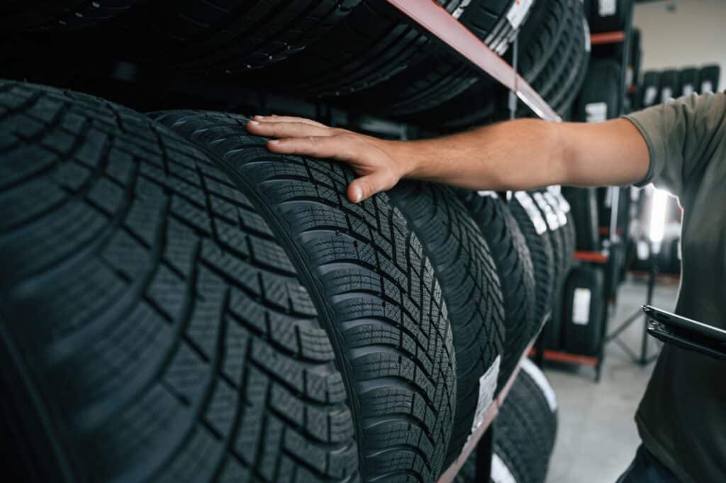 A person examines a row of stacked tires in a store or warehouse. Their hand rests on one of the tires to feel its tread pattern. The background shows multiple shelves filled with more tires.