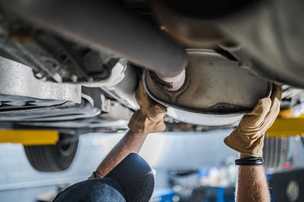 A mechanic wearing beige gloves and a blue cap works on the underside of a vehicle, focusing on an exhaust component. The background is slightly blurred, featuring a workshop setting with tools and equipment scattered.